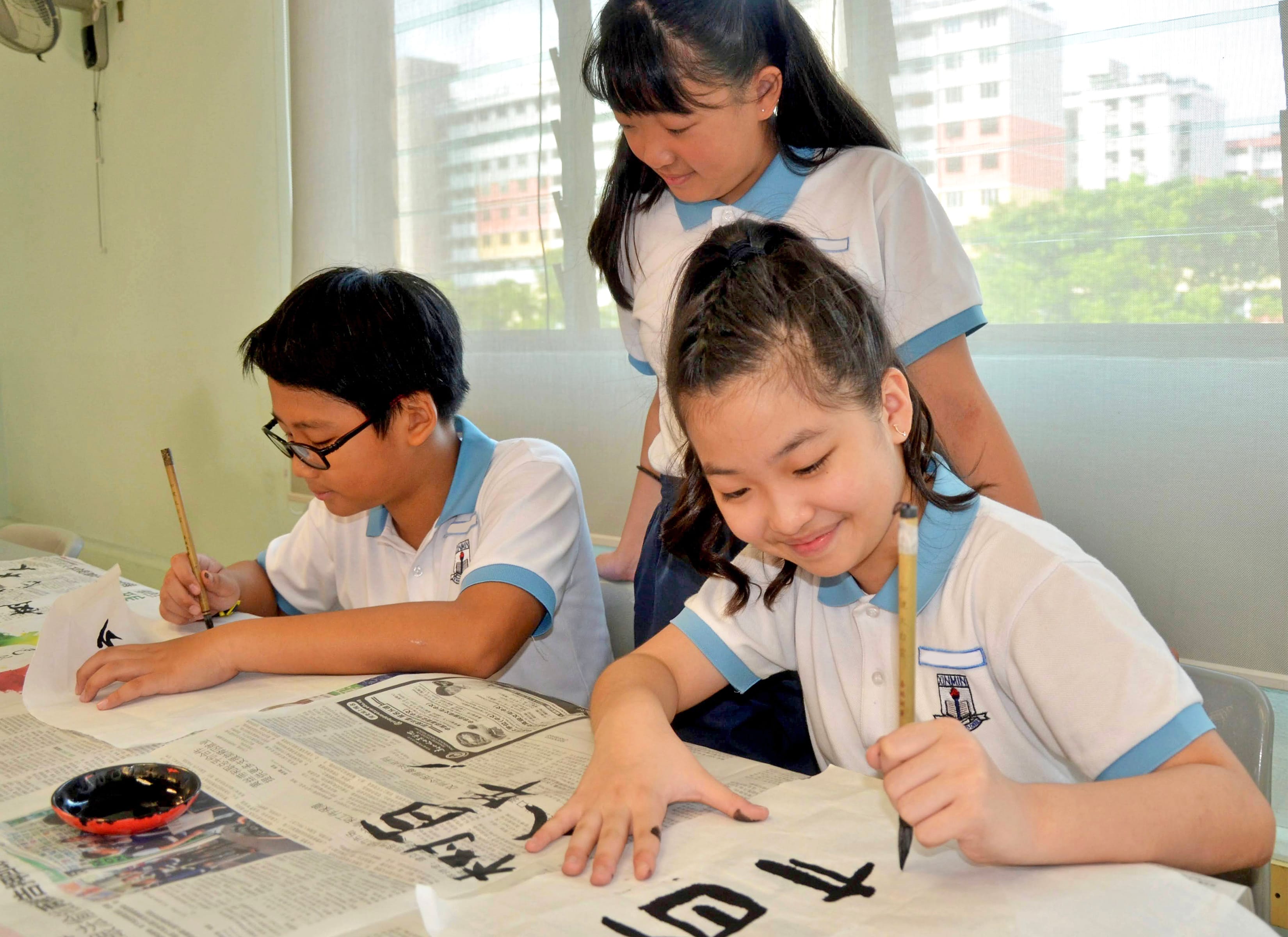 Three students in school uniforms practice Chinese calligraphy with brushes on paper at a table.